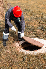 A worker in a hard hat lifts a manhole cover on a septic well. Inspection and maintenance of sewerage systems in rural areas