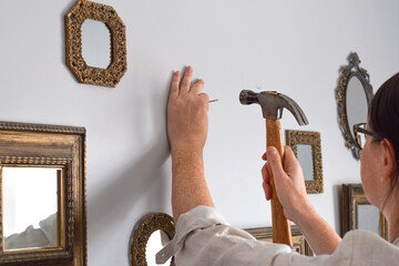 Closeup woman is putting nail in a wall with a hammer to hang mirrors in a gallery style