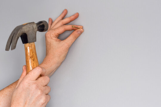 Closeup woman's hands putting nail in empty grey wall with a hammer
