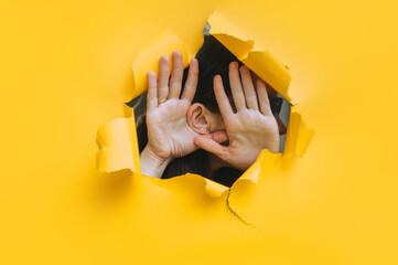 Close-up of a woman's ear and two hand through a torn hole in the paper. Yellow background, copy...