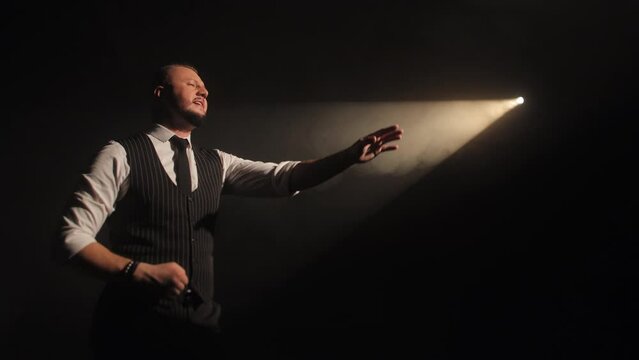 Actor in elegant suit and tie performing in contrast lighting and expressing personal emotions with hand gesture. Creative man singing beautiful song about love on scene in dramatic atmosphere.