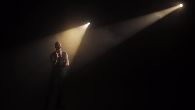 Handsome opera singer dressed in vest performing on stage in crossing of bright spotlights and making expressive hands gestures. Male musician acting and attracting attention of public by lyric song.