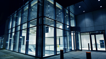 Night view on the ground floor of modern office building with big glass windows and entrance. Street reflection.