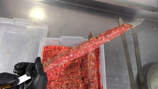 Chef sticks forcemeat on skewer and forming it for cooking lula kebab on grill at commercial kitchen. Close-up hands in black gloves
