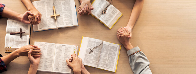 Cropped image of group of people praying together while holding hand on holy bible book at wooden...