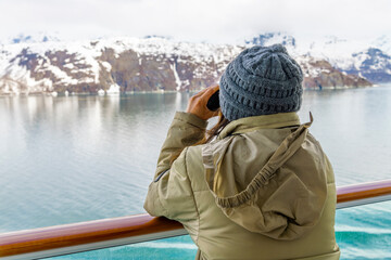 A young woman with binoculars views the snow covered mountains and glaciers from a cruise ship balcony at Glacier Bay National Park and Reserve, Alaska USA.	