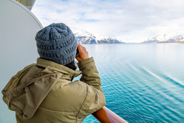 A young woman with binoculars views the snow covered mountains and glaciers from a cruise ship balcony at Glacier Bay National Park and Reserve, Alaska USA.	