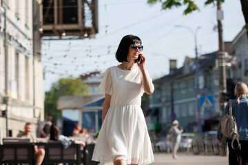 Romantic young brunette woman with short hair wears mini white dress and sunglasses, talking on cellphone, walking on a crowded city street in the summertime. Girl communicates by smartphone outdoors