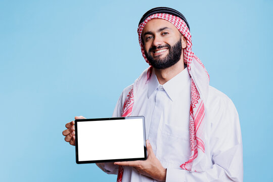 Smiling arab man presenting digital tablet empty screen for app presentation and looking at camera with cheerful expression. Arab in traditional clothes showing blank touchschreen in horizontal mode