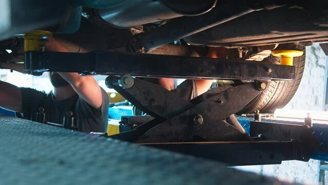 Two mechanics smilingly work on the chassis of a car, maneuvering around a lift in a noisy auto repair shop.