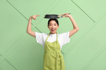 Portrait of young Asian woman pointing at frying pans on her head against green background