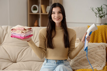 Pretty young woman with modern garment steamer and stack of clean clothes at home