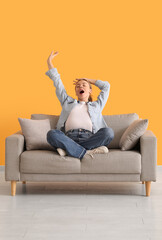 Beautiful young woman sitting on comfortable sofa and yawning near yellow wall