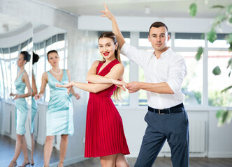 Fototapeta premium Expressive young adult woman in elegant vibrant red dress dancing passionate bachata with male partner during dance class for adults led by female instructor..