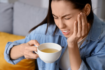 Young woman with cup of tea suffering from toothache at home, closeup