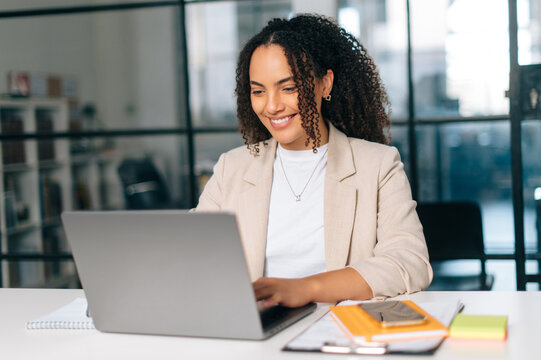 Amazing curly haired hispanic or brazilian young woman in stylish elegant clothes, office employee, small business owner, working on a laptop while sitting at her workplace in the modern office, smile