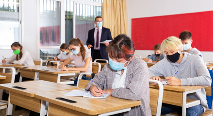 Teacher man in protective mask helping students during lesson in schoolroom
