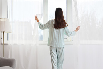 Young woman opening light curtains in living room. Back view