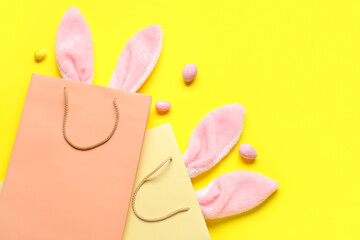 Shopping bags with fluffy bunny ears and Easter eggs on yellow background