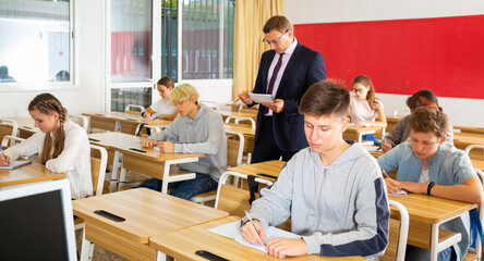Focused teenage students studying in classroom with teacher, writing lectures in workbooks