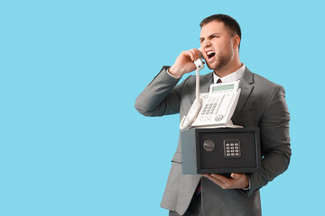 Angry young man with safe box talking by phone on blue background