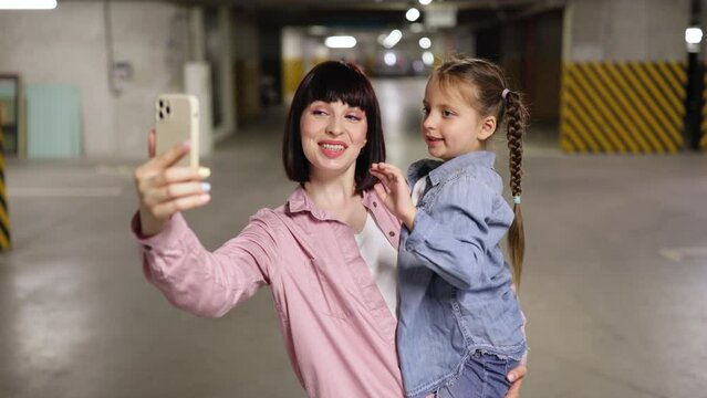 Caucasian mother holding preschool daughter in hands making selfie with smartphone in underground parking lot. Happy family in casual clothes having video call using cell phone before exciting trip.