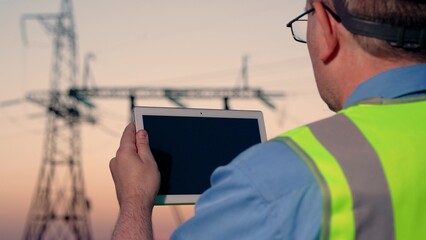 Power engineer in protective white helmet checks power line online using computer tablet, remote...