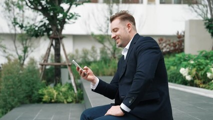 Smart caucasian businessman looking at smart phone and checking report. Happy investor smiling while talking or calling to manager about marketing plan while sitting at eco park or urban city. Urbane. - Powered by Adobe