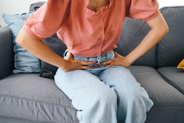 Young woman holding her stomach while sitting on the sofa. Stomach issues and problems concept 