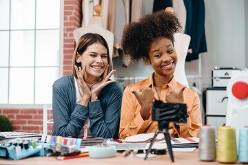 Two women are smiling and waving at the camera. They are sitting at a desk with online live to sale the product. Scene is friendly and lighthearted
