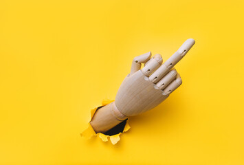 Middle finger of a rsght wooden hand, offensive gesture. Torn hole in yellow paper. Fuck you concept. Aggressive reaction of artificial intelligence or robot.