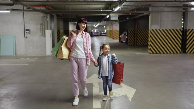 Caucasian mother and little daughter, in casual clothes, walking at underground parking lot. Young woman with little child holding colorful bags with clothes bought from shopping mall.
