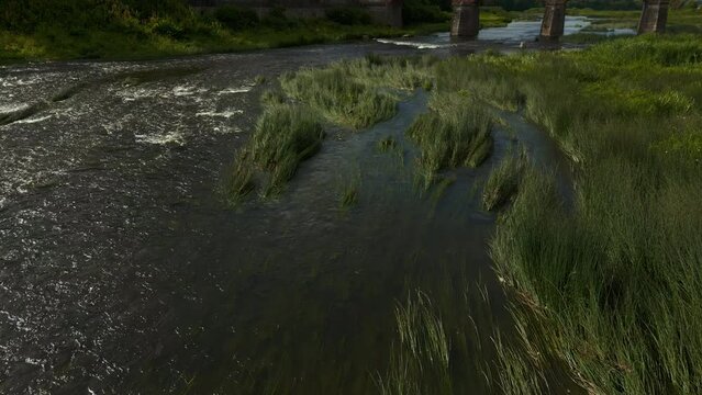 Hern aerial view: Water flowing through grassy field with bridge in natural landscape