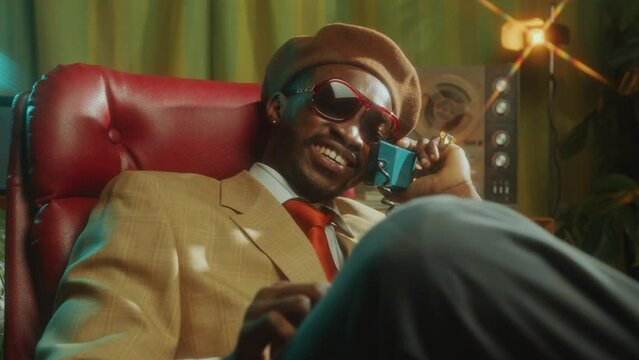 Smiling African-American man in retro outfit and sunglasses sitting in old-fashioned studio, speaking on vintage landline phone and snapping fingers during conversation