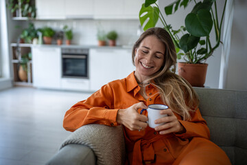 Pleased dreamy middle aged woman sits on couch relax at home holding cup coffee. Joyful female smiling thinking about happy moments, stress free, good mood, wellbeing enjoy weekend time in living room - obrazy, fototapety, plakaty