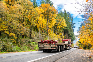 Industrial red big rig semi truck transporting empty flat bed semi trailer with rolled up tarp on the back running on the autumn narrow road with yellow trees on the hills