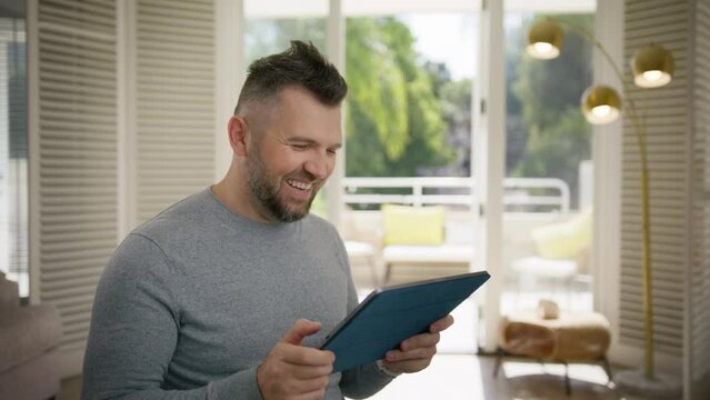 Happily smiling man looking at tablet screen and laughing out loud. Overjoyed middle age male browsing comic fun video content online on tablet. People using modern technology for leisure at home 4K