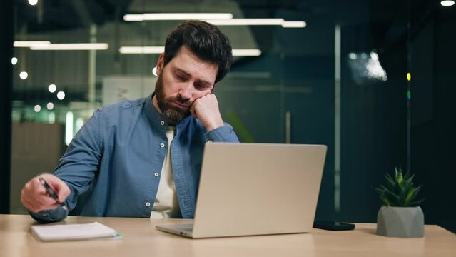 Unhappy male accountant typing on portable computer and finishing job while feeling tired. Sleepy man in informal attire yawning and covering mouth while waiting for end of boring working day.