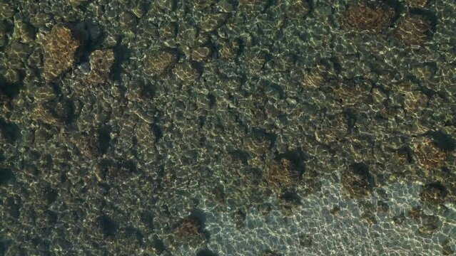 Drone top down bird's eye view of shimmering light ripples dancing across rocky boulders below water