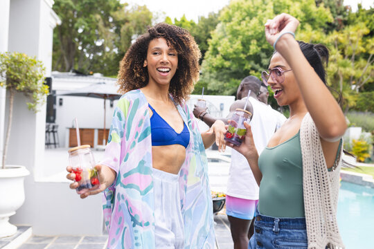 Biracial women enjoy a poolside party, one holding a drink and fruit