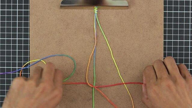 crafting a vibrant rainbow macrame bracelet with three threads, on a clipboard.