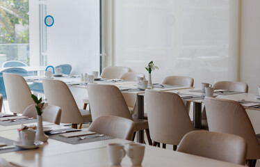 a dining area bathed in natural light. Tables are neatly arranged, each set with white dishes and cups. Beige chairs complement the setting. Large windows illuminate the space, enhancing the relaxed