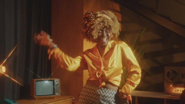 Young vibey Black woman in stylish look enjoying music and performing disco dance in retro studio with warm light, reel-to-reel tape player and old TV. Zoom Shot