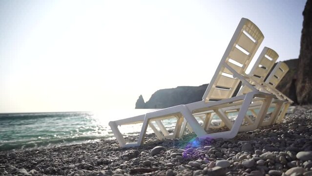 Rows of empty white plastic chair and sunbathing beds on beach near sea at sunset. Relaxation area for chilling out on summer. Beach Chairs and sun loungers outdoors. Summer Vacation resort concept.