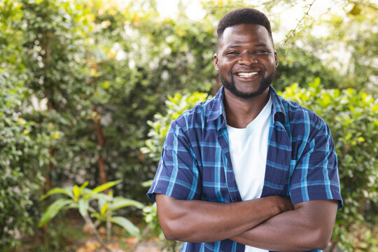 African American man smiles with arms crossed, wearing a blue plaid shirt, with copy space, in a gar