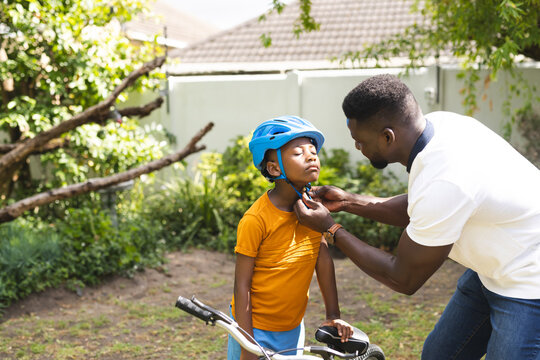 African American father secures a helmet on a young son's head, both outdoors in a backyard