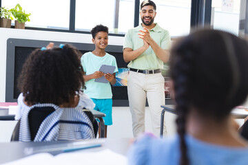 Young Asian male teacher stands applauding a biracial boy holding a tablet in a classroom in school
