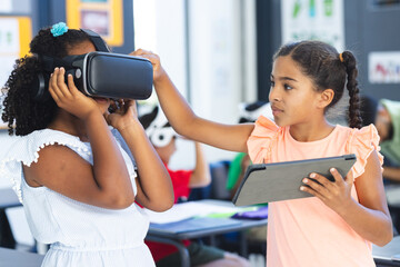 Biracial girl with a tablet assists another using VR headset in a classroom at school
