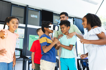 Young Asian male teacher assists a biracial boy with virtual reality headset as other children watch