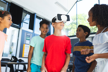 Biracial children are gathered around a boy using a virtual reality headset in a classroom at school